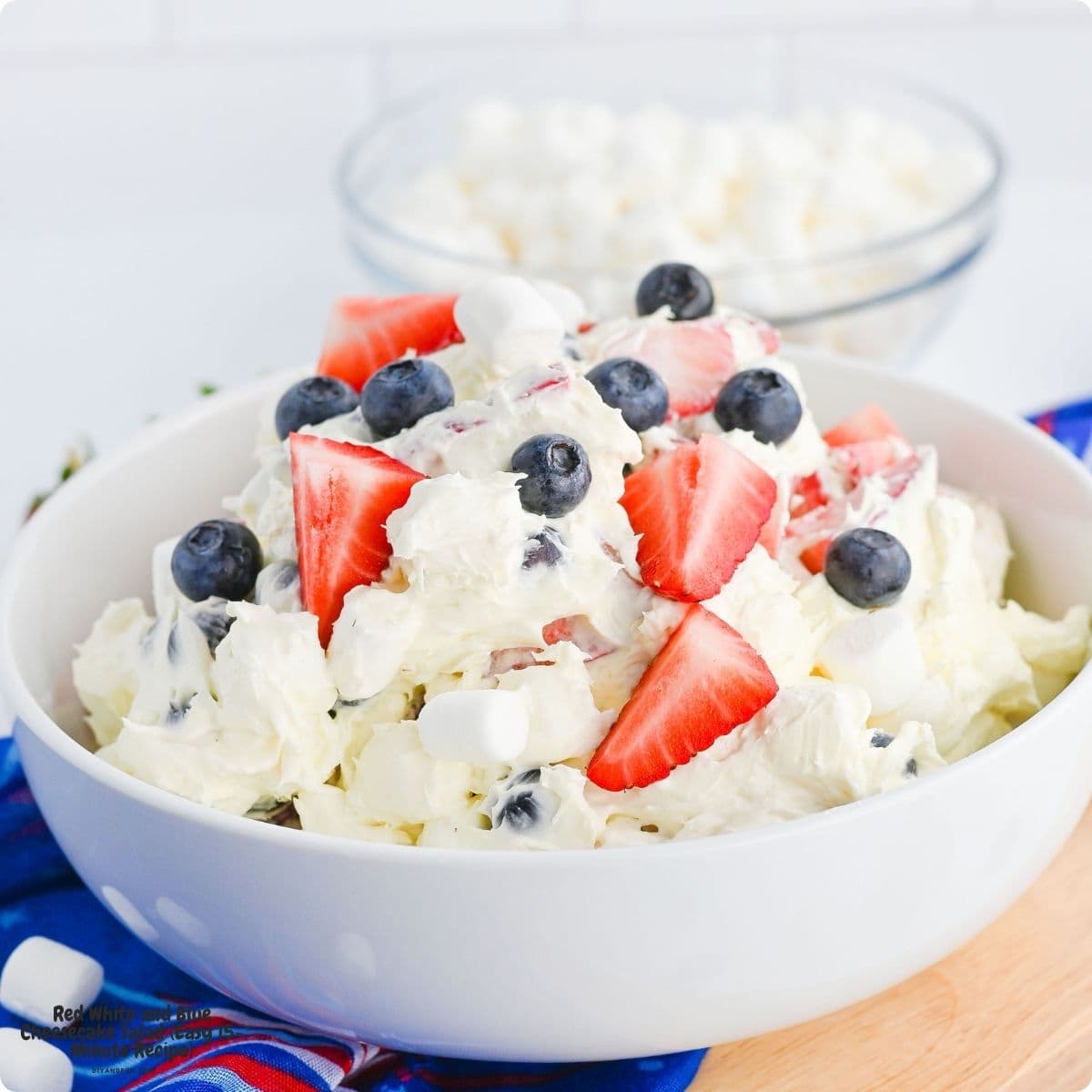 Red White and Blue Cheesecake Salad is an Easy 15-Minute Recipe for the ultimate creamy dessert summer made with fresh berries. Close up of fluffy cheesecake salad with mini marshmallows and quartered strawberries on a spoon